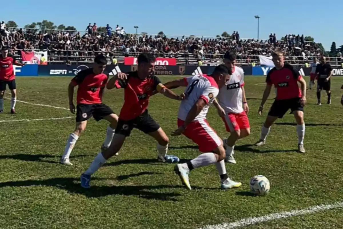 Jugadores de fútbol compiten por el balón en un partido con público en las gradas.