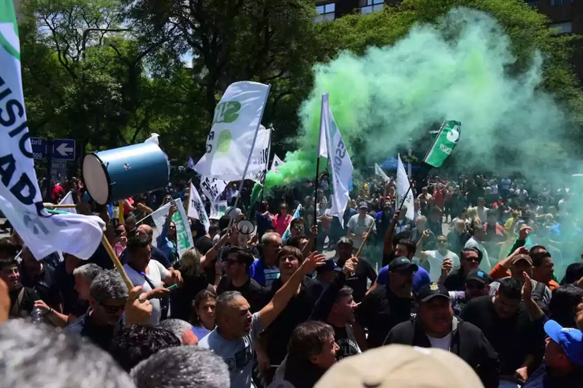 Una multitud de personas participa en una manifestación al aire libre con banderas y humo verde en el aire.