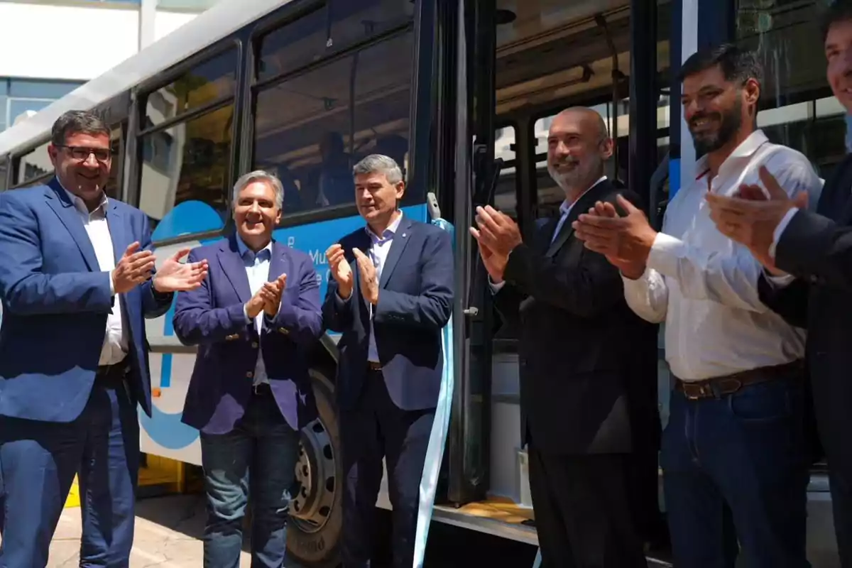 Un grupo de hombres aplaude frente a un autobús durante un evento al aire libre.