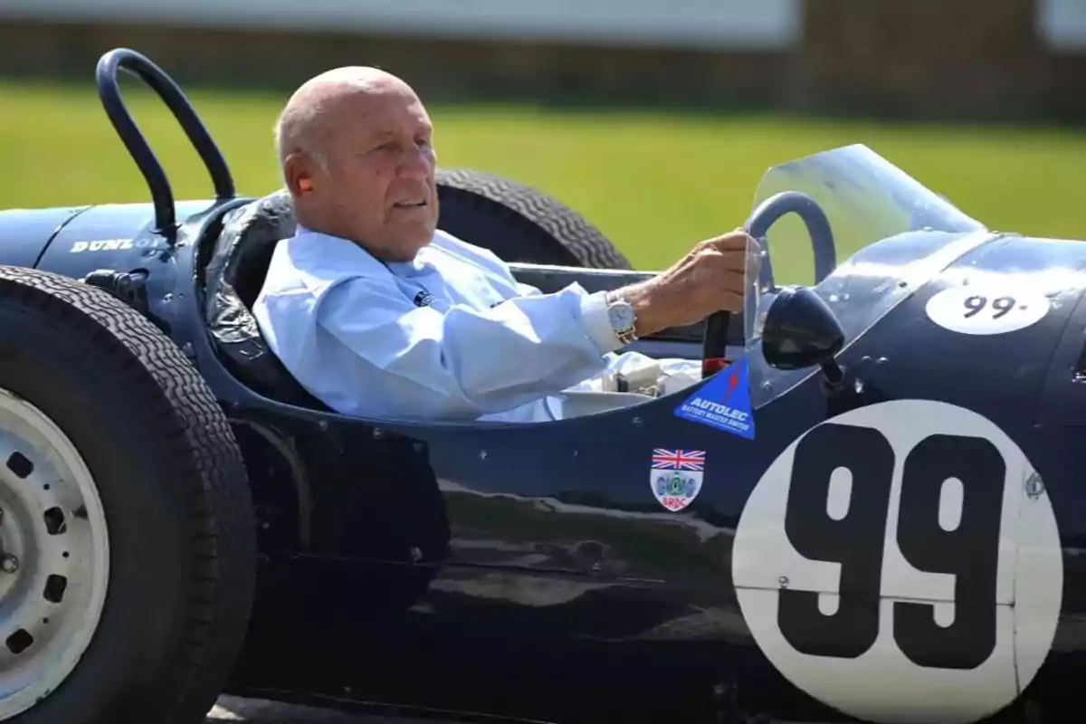 Un hombre mayor conduciendo un auto de carreras clásico con el número 99 en un entorno al aire libre.