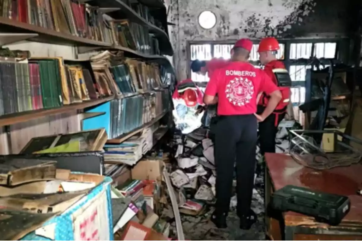 Bomberos inspeccionan una biblioteca dañada por un incendio con estantes llenos de libros quemados y escombros en el suelo.