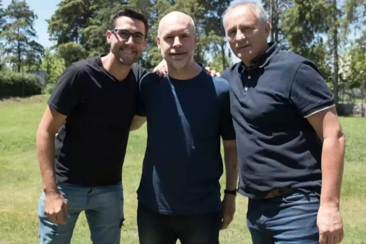 Tres hombres posan juntos al aire libre en un día soleado con árboles al fondo.