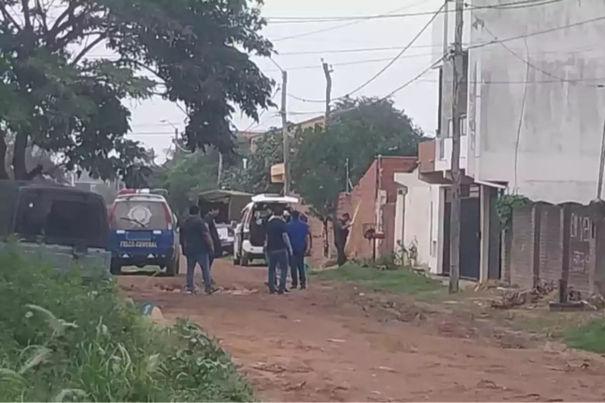 Un grupo de personas se encuentra en una calle de tierra junto a vehículos policiales y una camioneta, rodeados de árboles y edificios.