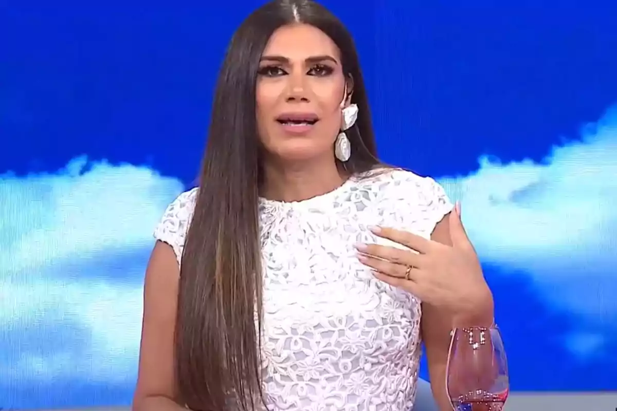 Mujer con cabello largo y liso usando un vestido blanco con detalles florales y grandes pendientes blancos, hablando en un set de televisión con fondo azul y nubes.