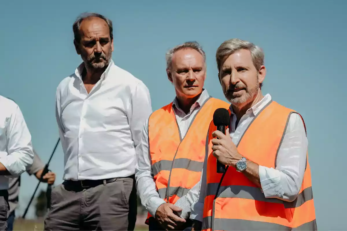 Tres hombres con chalecos de seguridad naranjas, uno de ellos hablando por un micrófono.