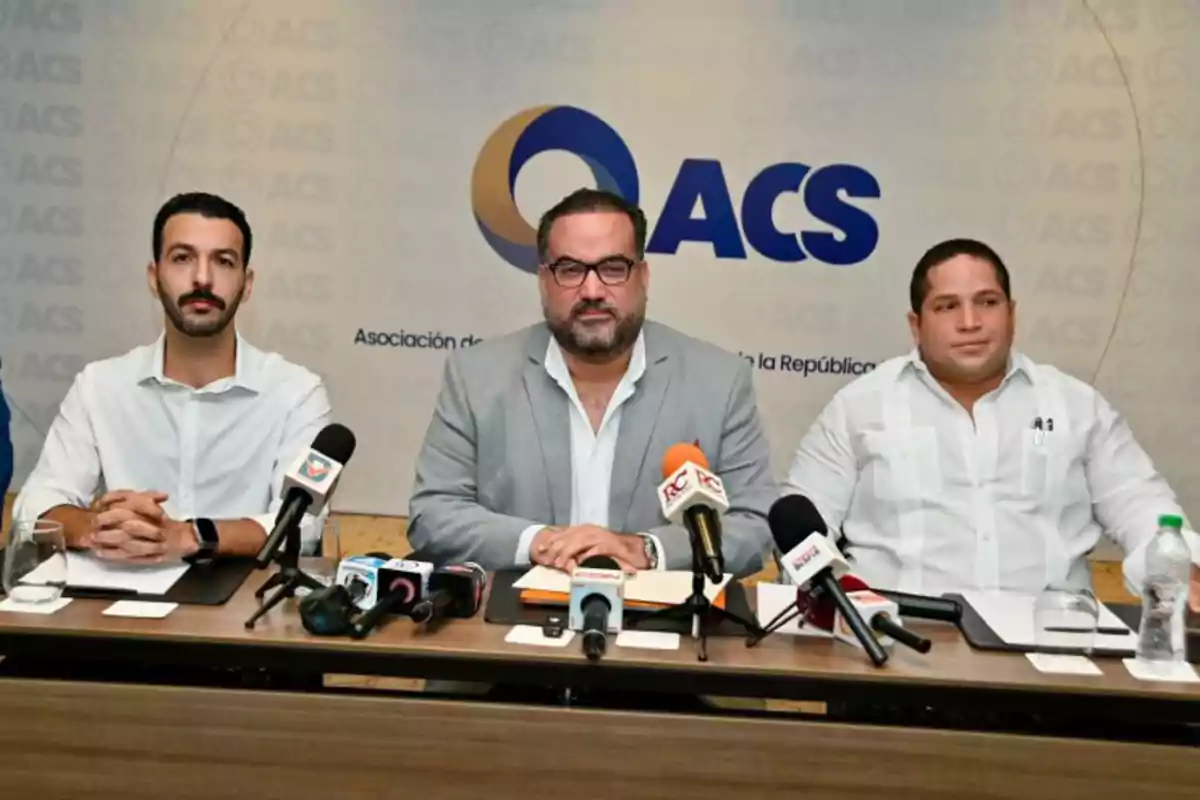 Tres hombres sentados en una mesa durante una conferencia de prensa con micrófonos frente a ellos y un fondo con el logo de ACS