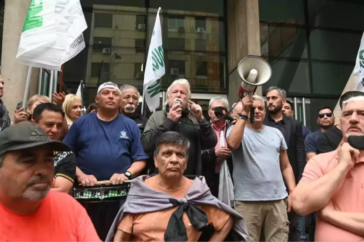 Un grupo de personas se encuentra reunido frente a un edificio, algunas sostienen banderas y una persona habla por un megáfono.