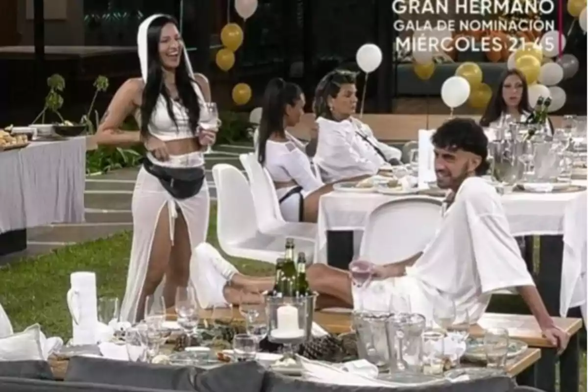 Un grupo de personas vestidas de blanco disfruta de una reunión al aire libre con mesas decoradas y globos en el fondo.