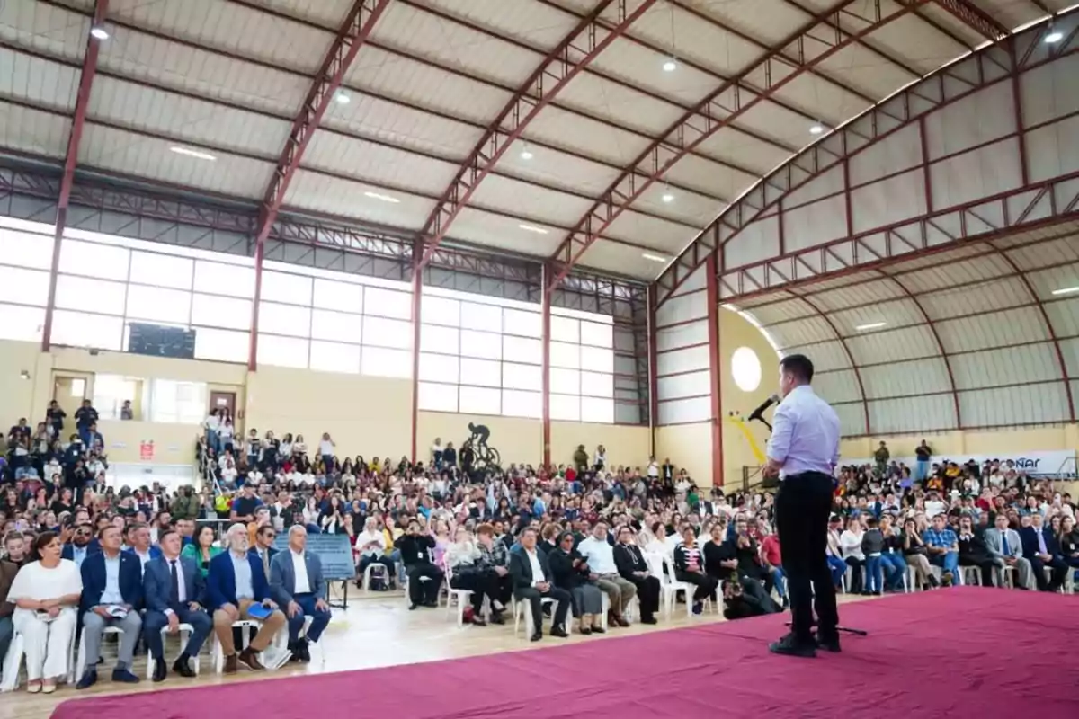 A person speaking into a microphone in front of a large audience in an indoor gym.