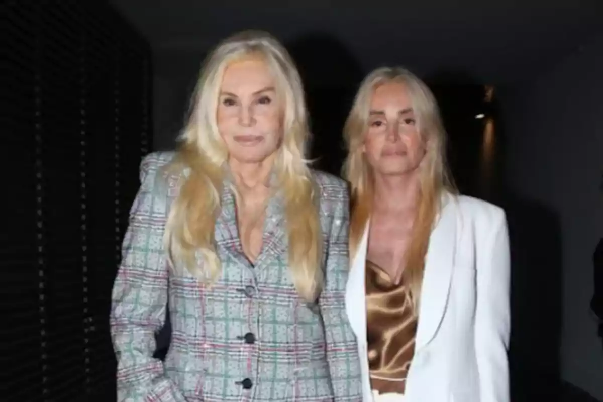 Dos mujeres con cabello rubio posan juntas, una lleva un traje a cuadros y la otra un conjunto blanco con blusa dorada.