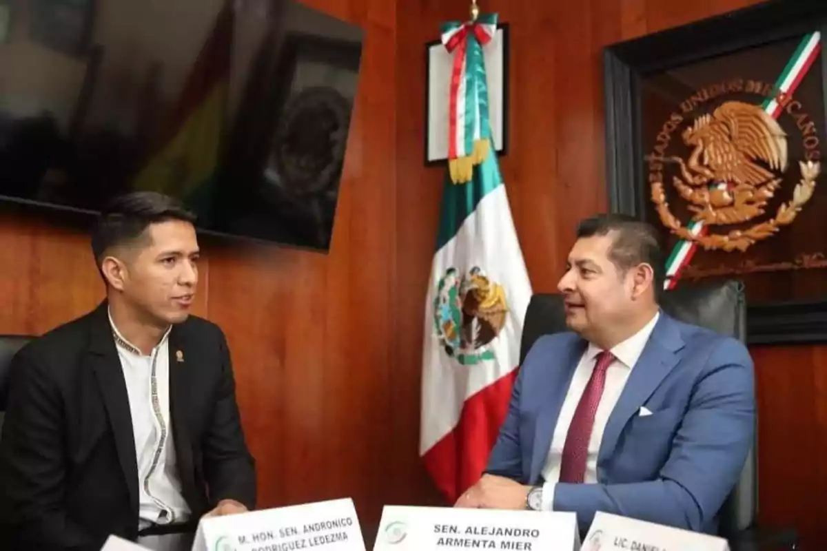 Dos hombres en traje conversan en una oficina con una bandera de México y un escudo en la pared.