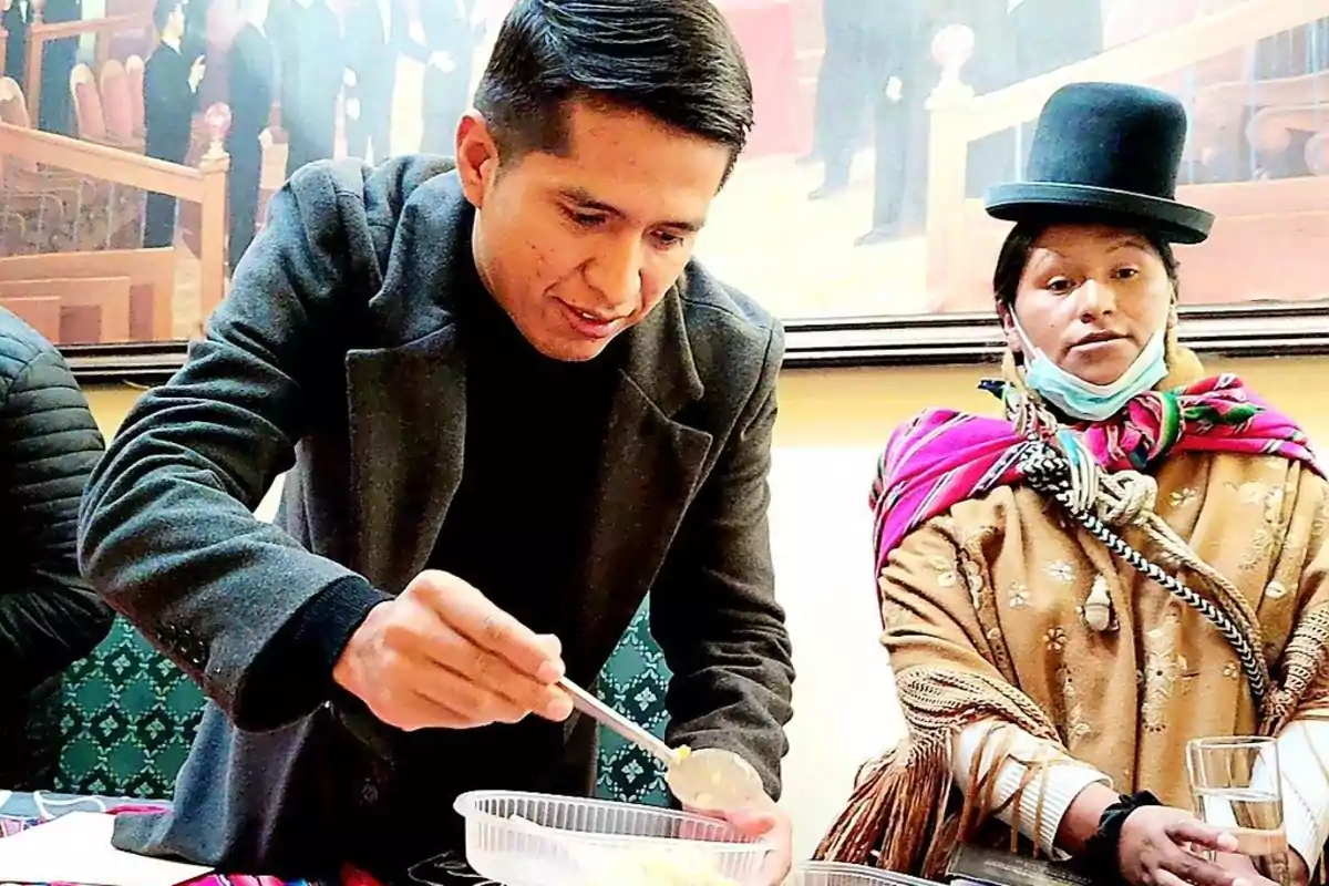 Un hombre sirviendo comida en un recipiente mientras una mujer con vestimenta tradicional observa.