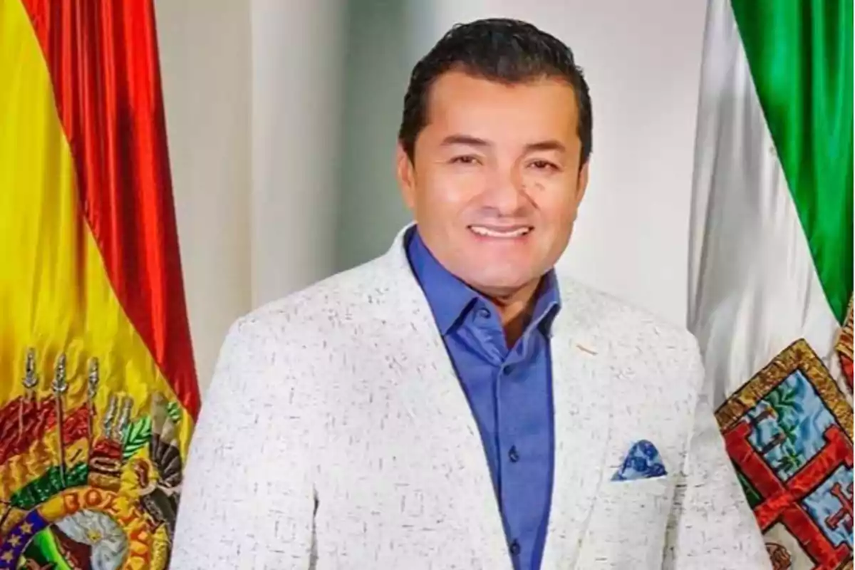 Un hombre con saco blanco y camisa azul está de pie frente a las banderas de Bolivia y México.