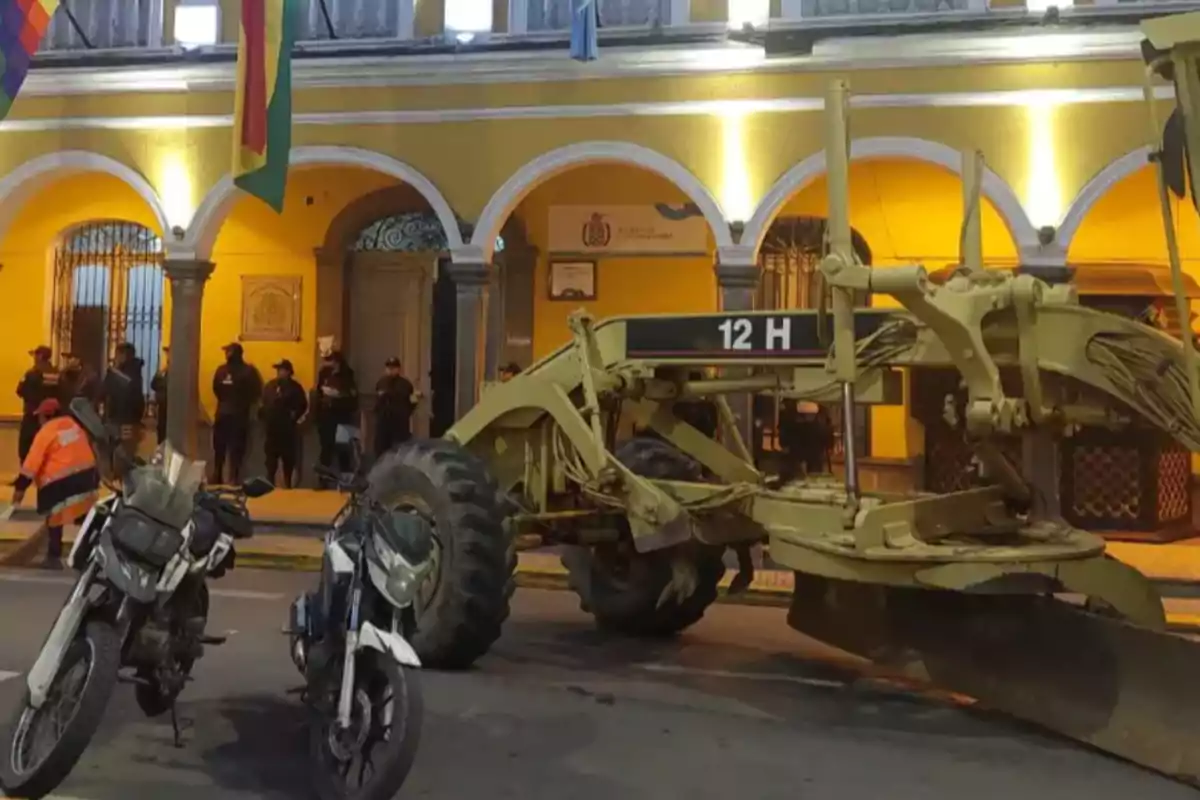 Una motoniveladora y dos motocicletas estacionadas frente a un edificio amarillo con arcos y banderas, mientras varias personas están de pie en el fondo.