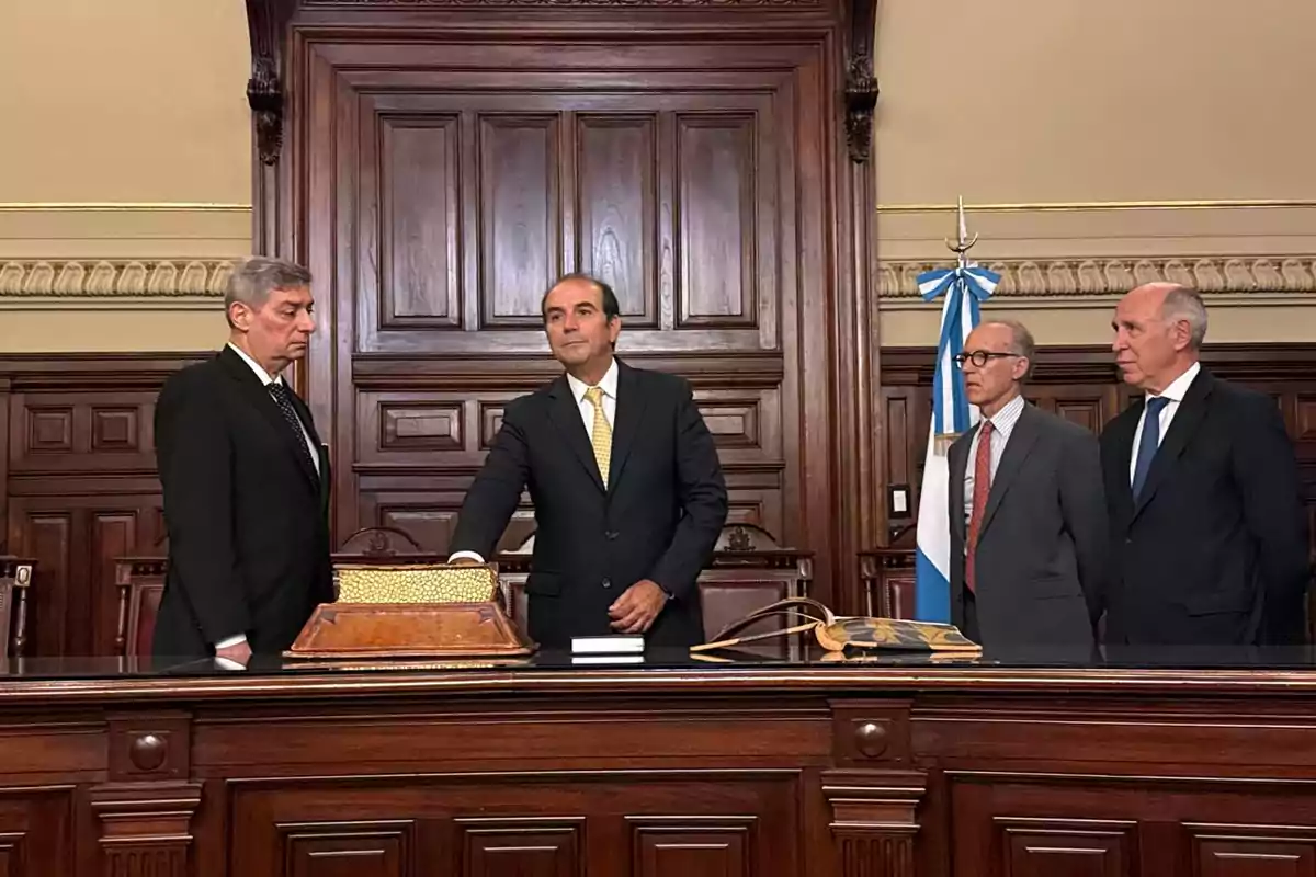 A group of men in formal suits is in a room with a carved wooden background and a blue and white flag.