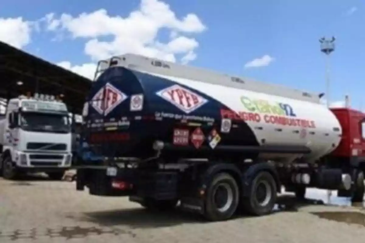 Un camión cisterna de combustible estacionado en una zona de carga con otros camiones alrededor y un cielo parcialmente nublado.