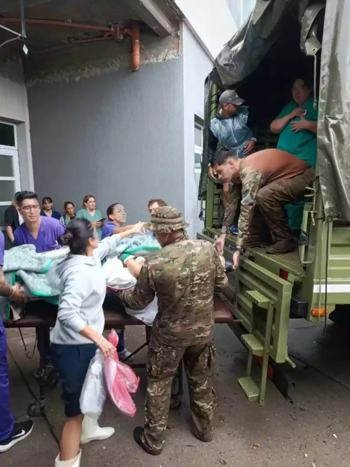 Un grupo de personas, incluyendo personal militar y médico, colabora para trasladar a un paciente en una camilla desde un camión militar hacia un edificio.