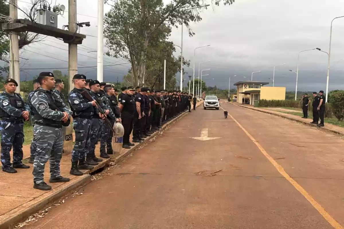 Un grupo de policías uniformados está alineado a lo largo de una carretera, con un vehículo y un perro en el centro de la escena.