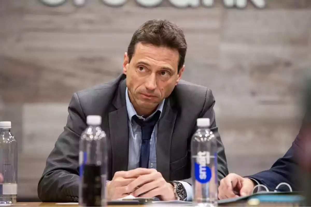 A man in a suit and tie is sitting at a table with bottles of water in front of him, looking thoughtfully to the side.