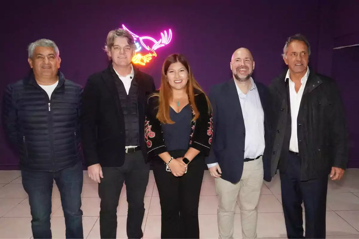 Cinco personas posan juntas frente a un fondo morado con un letrero de neón en forma de ave.