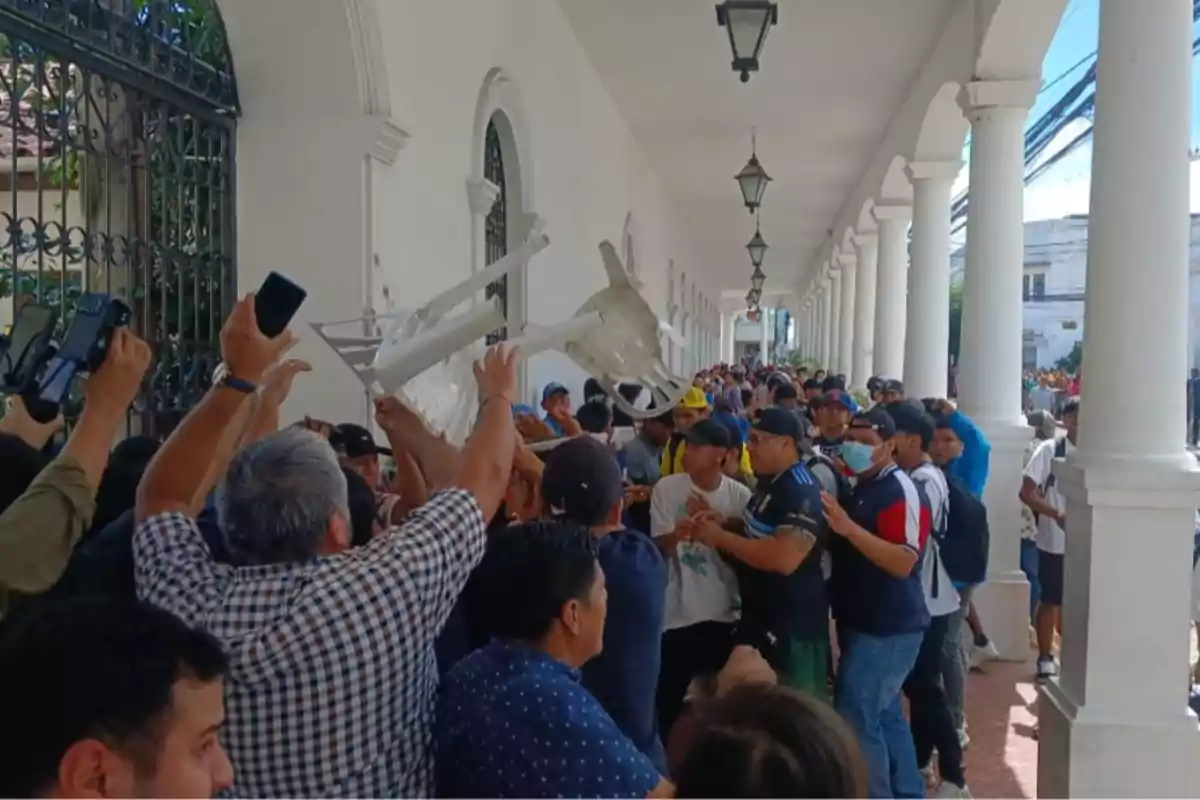A group of people is gathered in a hallway with white columns, some are raising a plastic chair in the air while others watch and take photos. A group of people is gathered in a hallway with white columns, some are raising a plastic chair in the air while others watch and take photos.