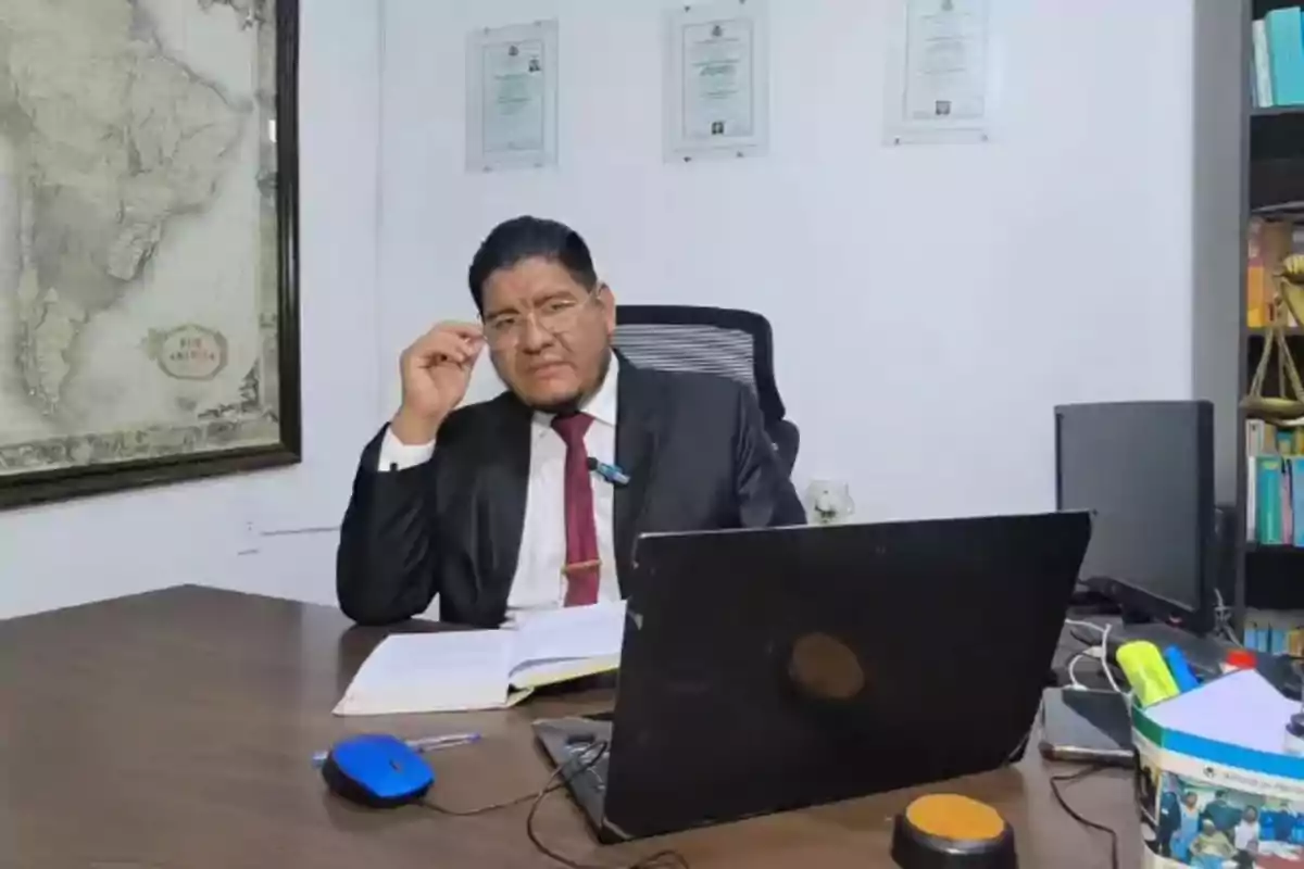 Un hombre con traje y corbata está sentado en un escritorio con una computadora portátil y documentos, en una oficina con un mapa en la pared y certificados enmarcados. Un hombre con traje y corbata está sentado en un escritorio con una computadora portátil y documentos, en una oficina con un mapa en la pared y certificados enmarcados.