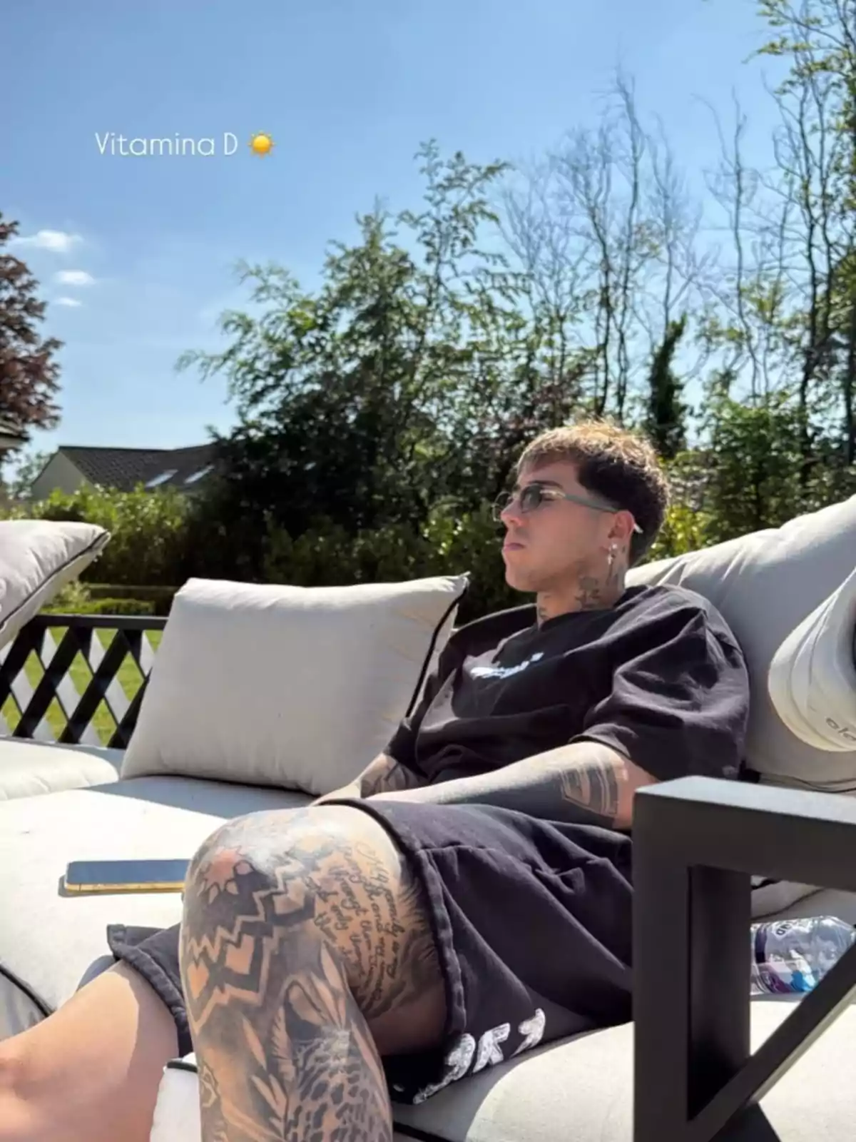 Una persona con tatuajes está sentada en un sofá al aire libre disfrutando del sol, rodeada de vegetación y con un cielo despejado. Una persona con tatuajes está sentada en un sofá al aire libre disfrutando del sol, rodeada de vegetación y con un cielo despejado.