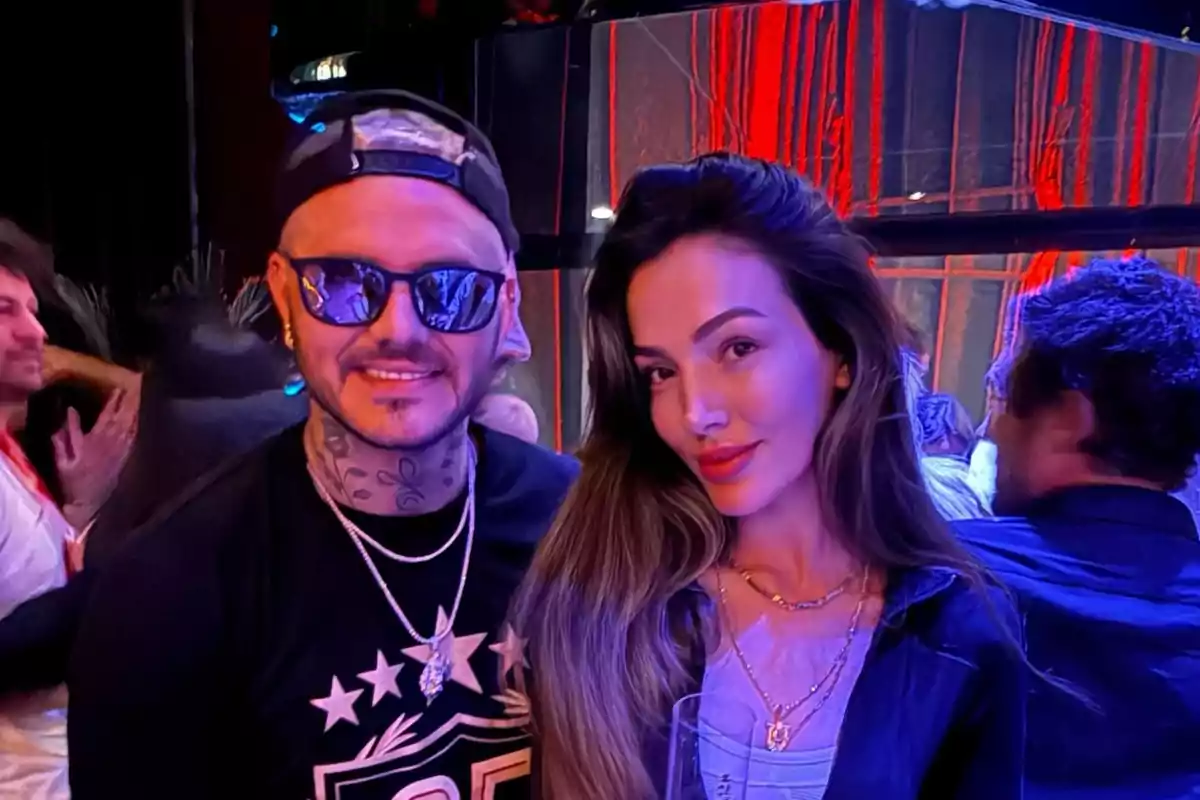Two people pose together in an environment lit with colorful lights, one of them is wearing sunglasses and a cap, while the other has long, loose hair.