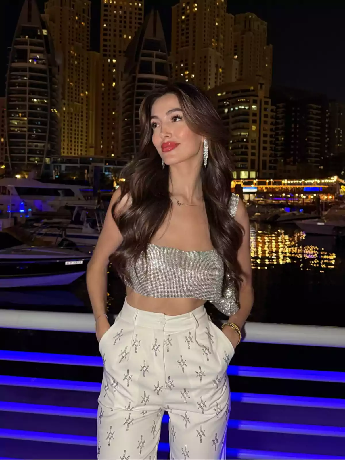 A woman in a shiny top and decorated white pants, posing in front of a harbor lit up at night with tall buildings in the background. A woman in a shiny top and decorated white pants, posing in front of a harbor lit up at night with tall buildings in the background.