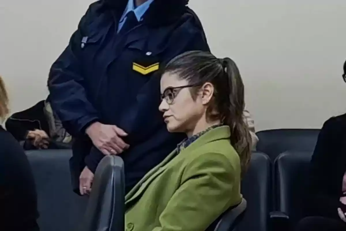 Mujer con gafas y saco verde sentada en una sala junto a otras personas con un oficial de pie detrás de ella