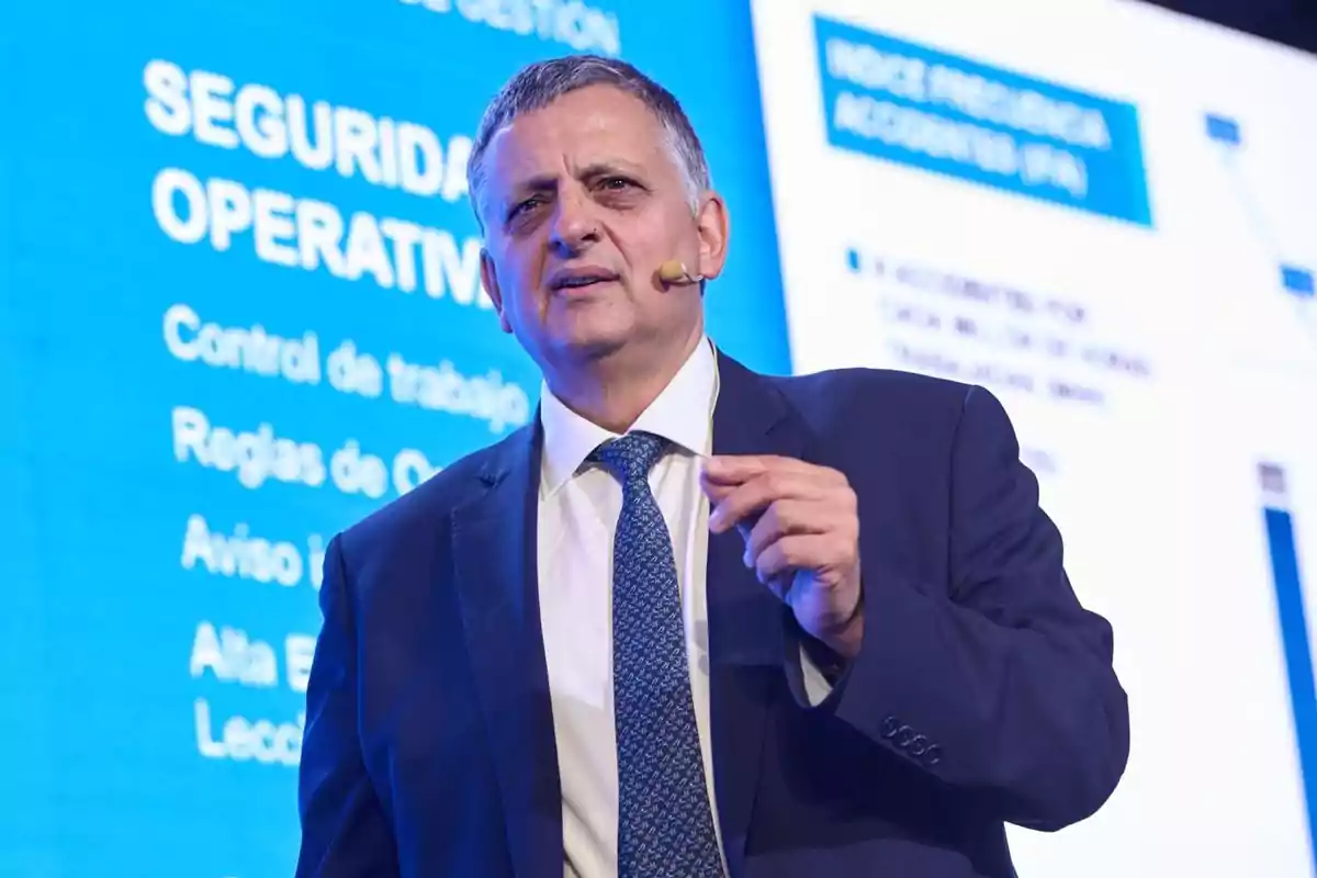 A man in a suit and tie speaks at a conference, with a blue screen in the background displaying text related to operational security.