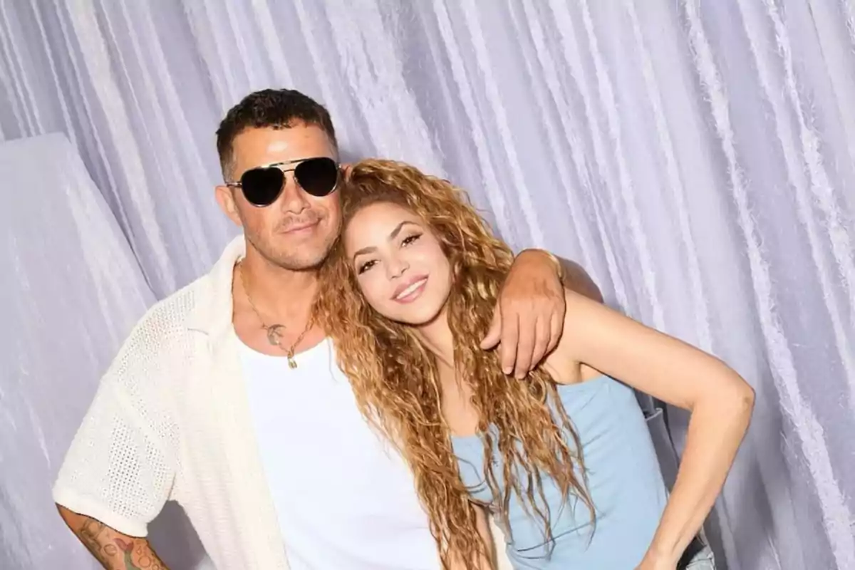 Dos personas posando juntas frente a un fondo claro, una de ellas lleva gafas de sol y la otra tiene el cabello rizado.