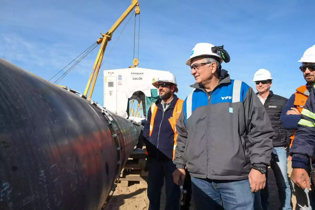 Several workers wearing helmets and vests watch the installation of a large gas pipeline at an outdoor construction site.