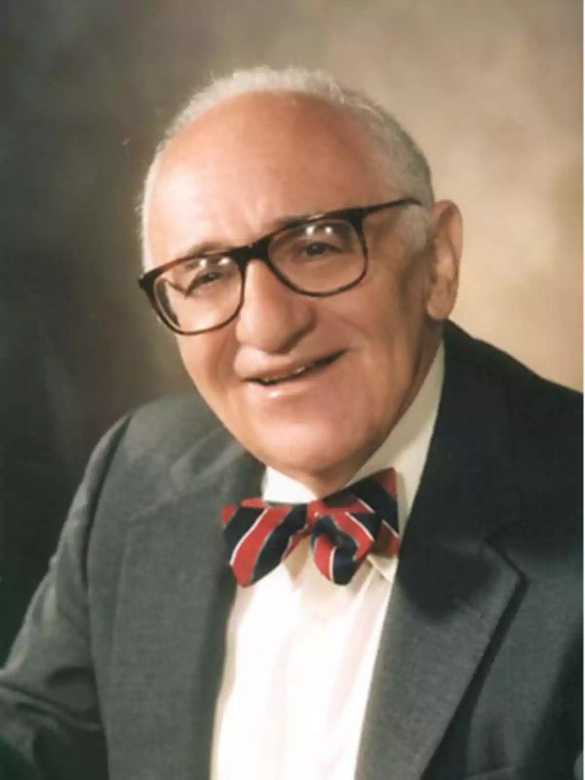 Older man with glasses, gray hair, dark suit, and a red-and-black striped bow tie, smiling in front of a neutral background Older man with glasses, gray hair, dark suit, and a red-and-black striped bow tie, smiling in front of a neutral background