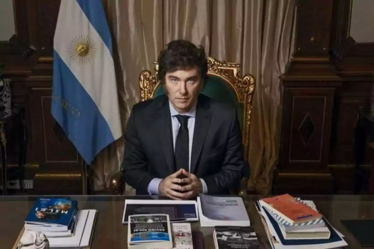 Man in a dark suit sitting at a desk with several books and documents, with an Argentine flag and beige curtains in the background