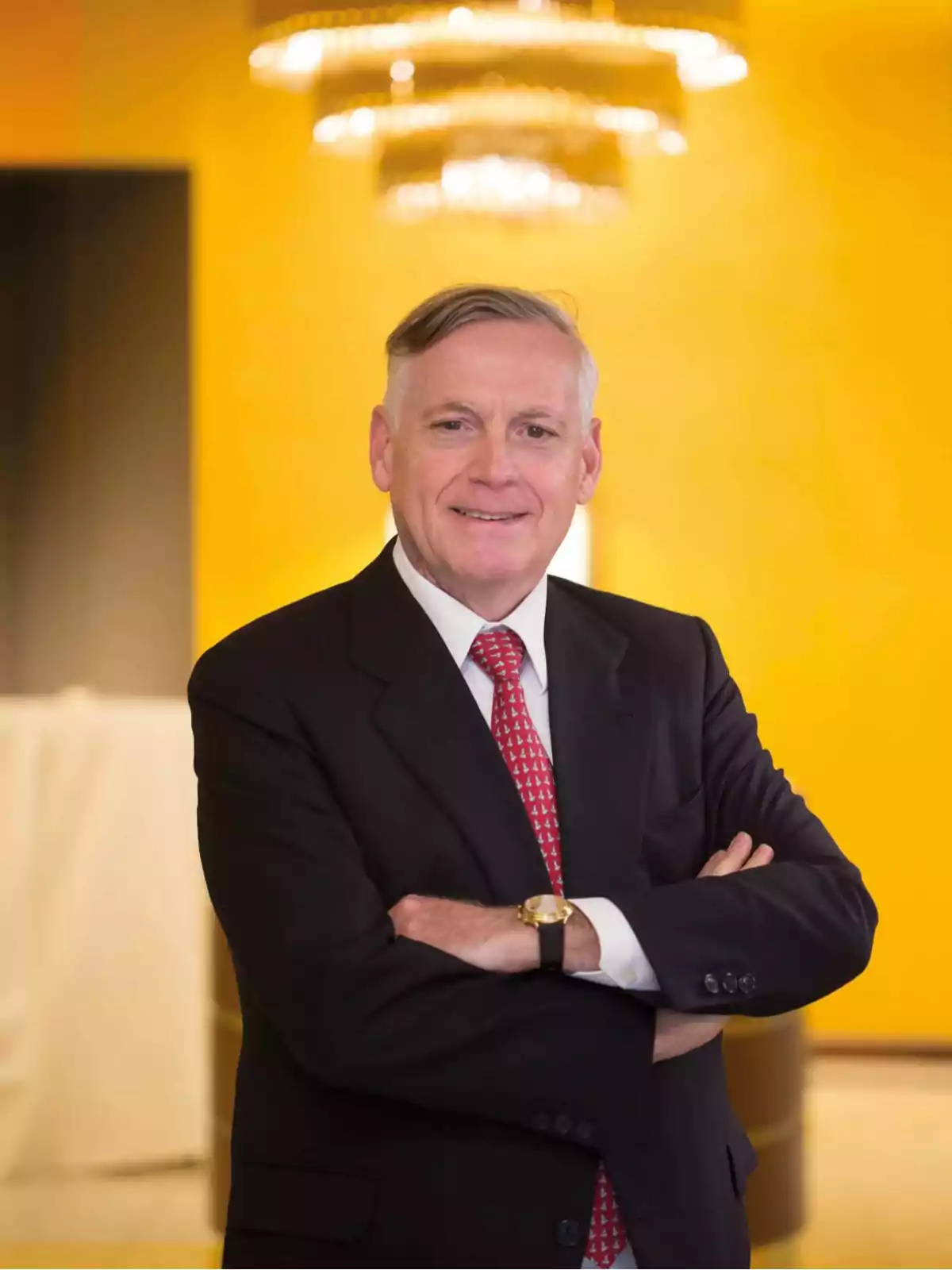 Gray-haired man in a dark suit and red tie posing with his arms crossed in front of a yellow wall and an elegant lamp in the background Gray-haired man in a dark suit and red tie posing with his arms crossed in front of a yellow wall and an elegant lamp in the background