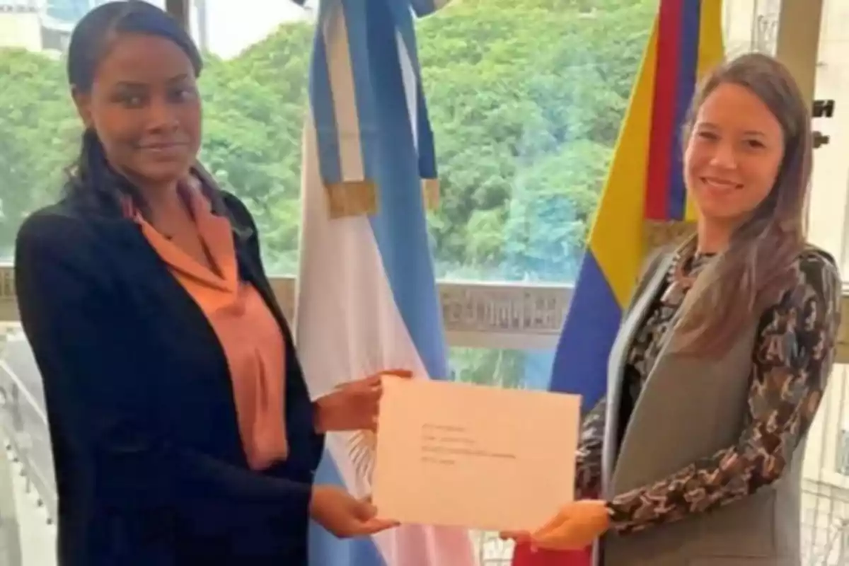 Two women smile while holding a document in front of the flags of Argentina and Colombia in an office setting with large windows and trees in the background.