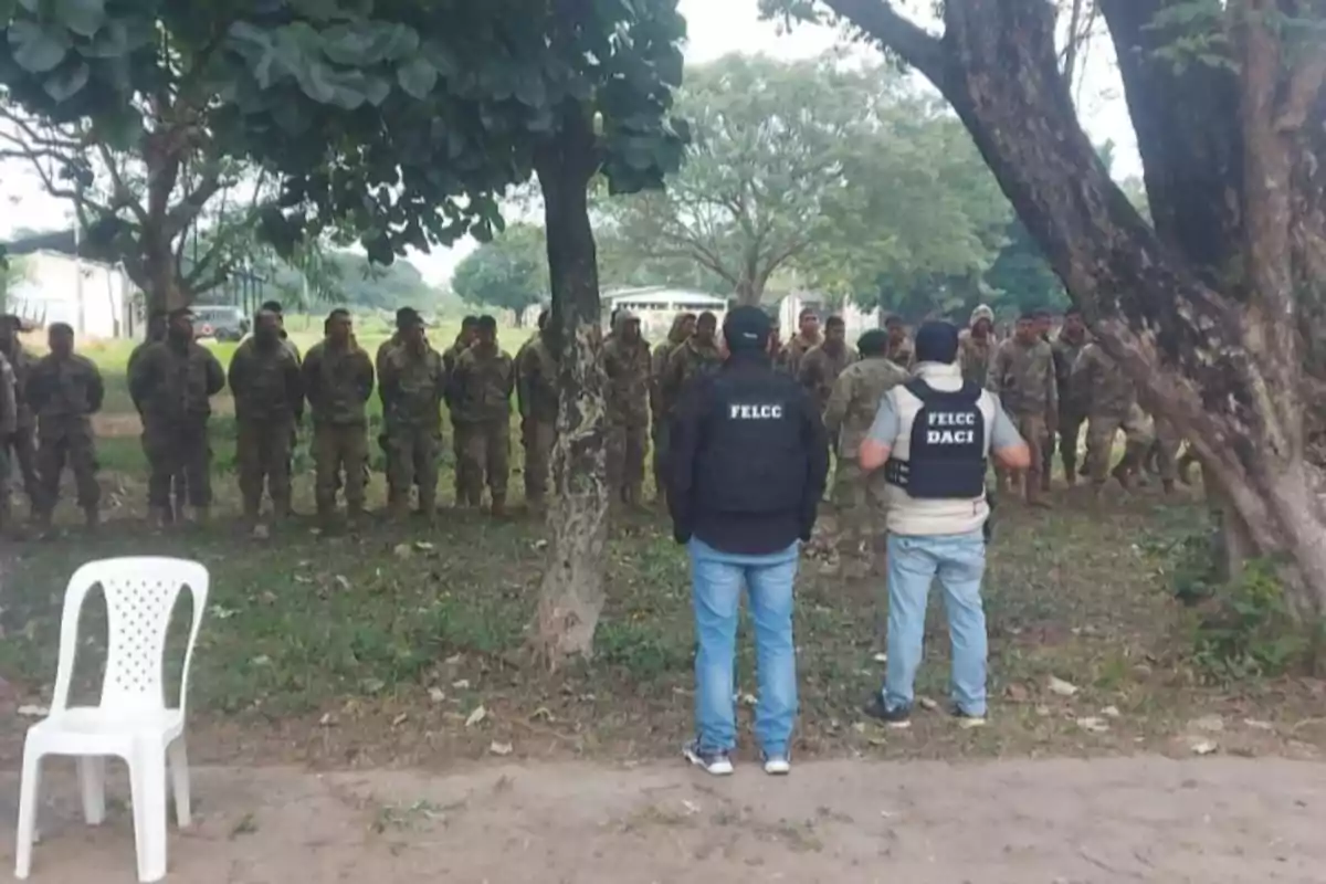 Un grupo de militares está formado en fila en un área verde mientras dos personas con chalecos de la FELCC observan de espaldas y una silla blanca de plástico está vacía en primer plano.