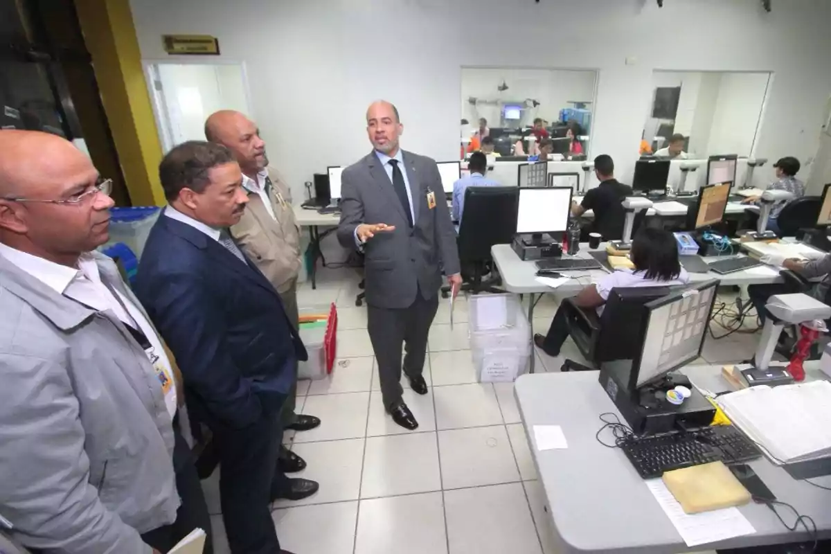 Un grupo de hombres de traje y chaqueta observa mientras un hombre de traje les explica algo en una oficina con varias personas trabajando en computadoras.