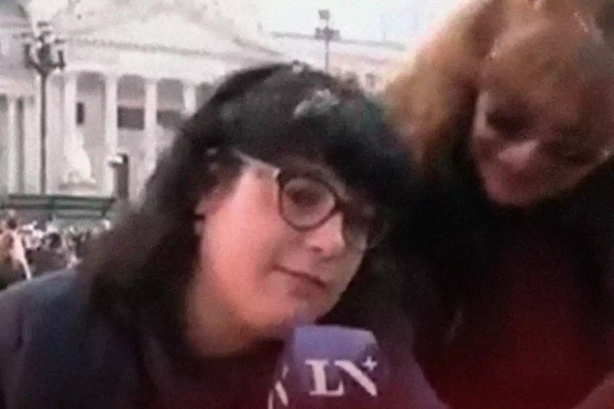 Two women appear in the foreground during a street interview, one of them is holding a microphone and wearing glasses, in the background there is a white building and a crowd.