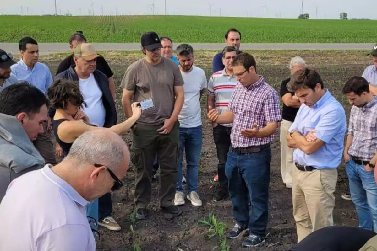 Un grupo de personas observa y conversa en un campo de cultivo mientras una mujer sostiene un micrófono y un hombre explica algo con las manos abiertas