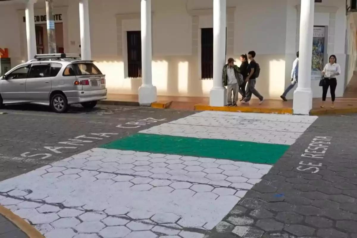 Un paso peatonal pintado con franjas blancas y una franja verde en el centro, junto a una acera donde caminan varias personas y un automóvil estacionado a la izquierda. Un paso peatonal pintado con franjas blancas y una franja verde en el centro, junto a una acera donde caminan varias personas y un automóvil estacionado a la izquierda.