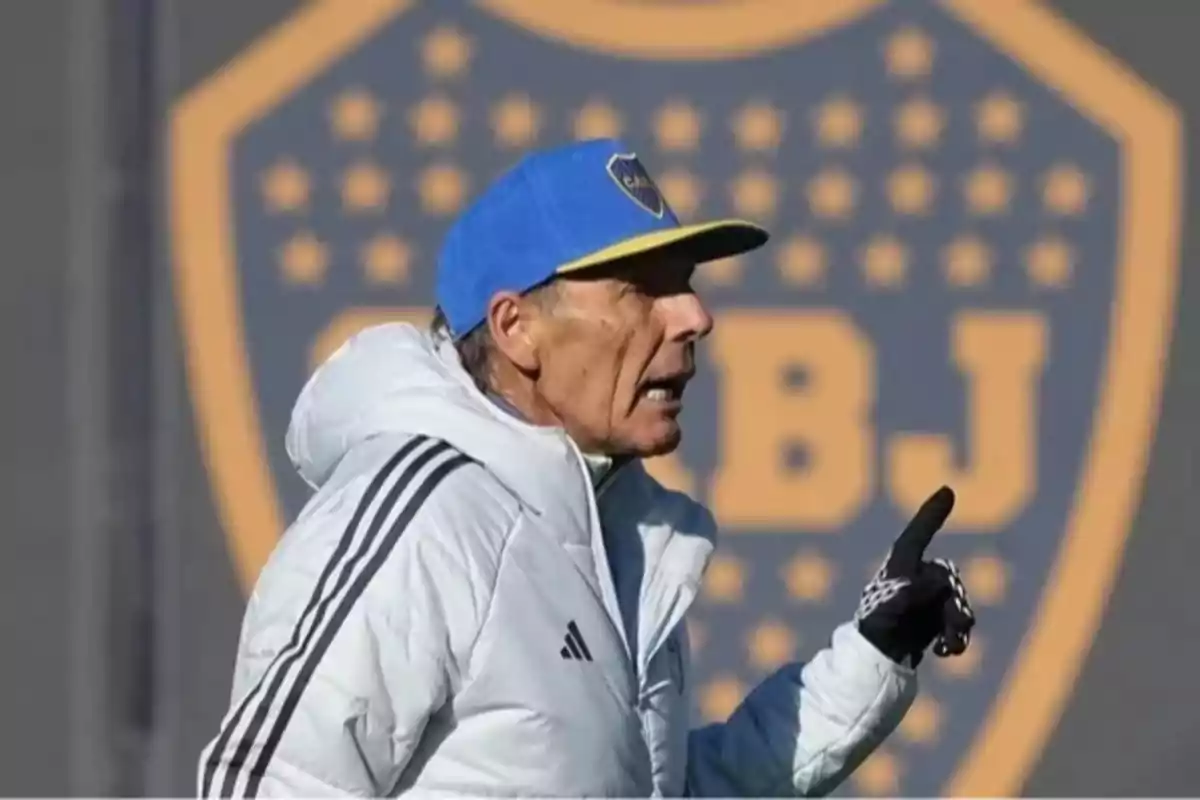 Entrenador de fútbol con gorra azul y campera blanca dando indicaciones frente a un escudo de Boca Juniors desenfocado de fondo