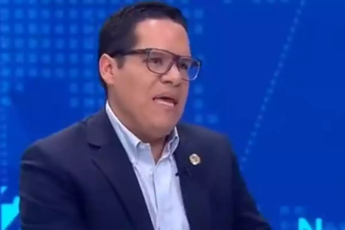 Man with glasses and a dark jacket speaking on a television set with a blue background Man with glasses and a dark jacket speaking on a television set with a blue background