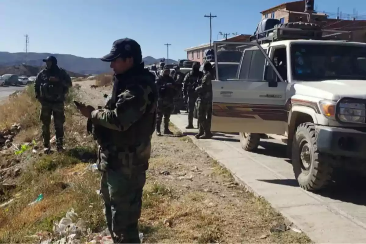 Varios policías con uniforme camuflado están de pie junto a una camioneta blanca estacionada en una calle rural durante el día Varios policías con uniforme camuflado están de pie junto a una camioneta blanca estacionada en una calle rural durante el día
