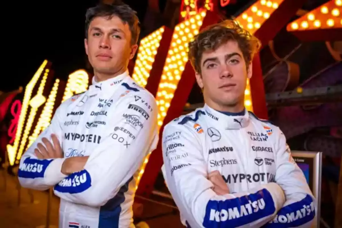 Dos pilotos de carreras con uniformes blancos posan con los brazos cruzados frente a un fondo iluminado.