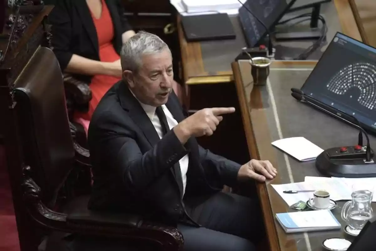 A man in a suit is sitting at a desk pointing with his finger while speaking, with a laptop and a cup of coffee on the table.
