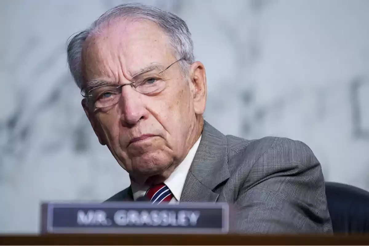 Hombre mayor de cabello canoso y gafas con expresión seria, usando traje y corbata, sentado frente a un letrero que dice MR GRASSLEY Hombre mayor de cabello canoso y gafas con expresión seria, usando traje y corbata, sentado frente a un letrero que dice MR GRASSLEY