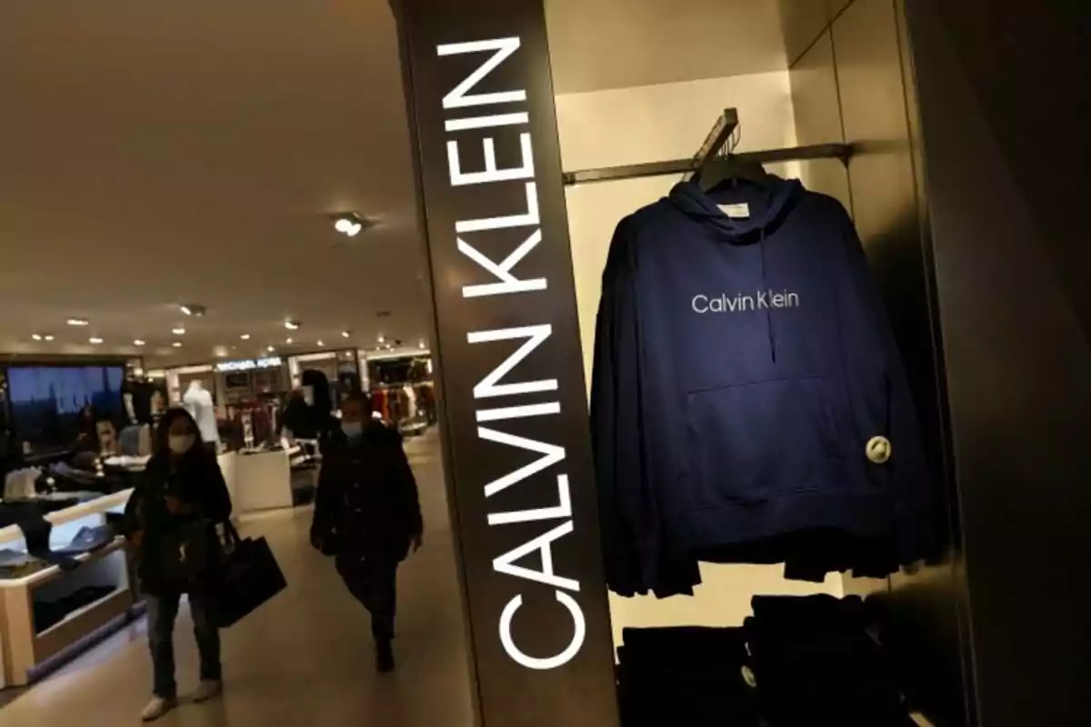 Una sudadera azul de Calvin Klein colgada en una tienda iluminada con personas caminando al fondo.