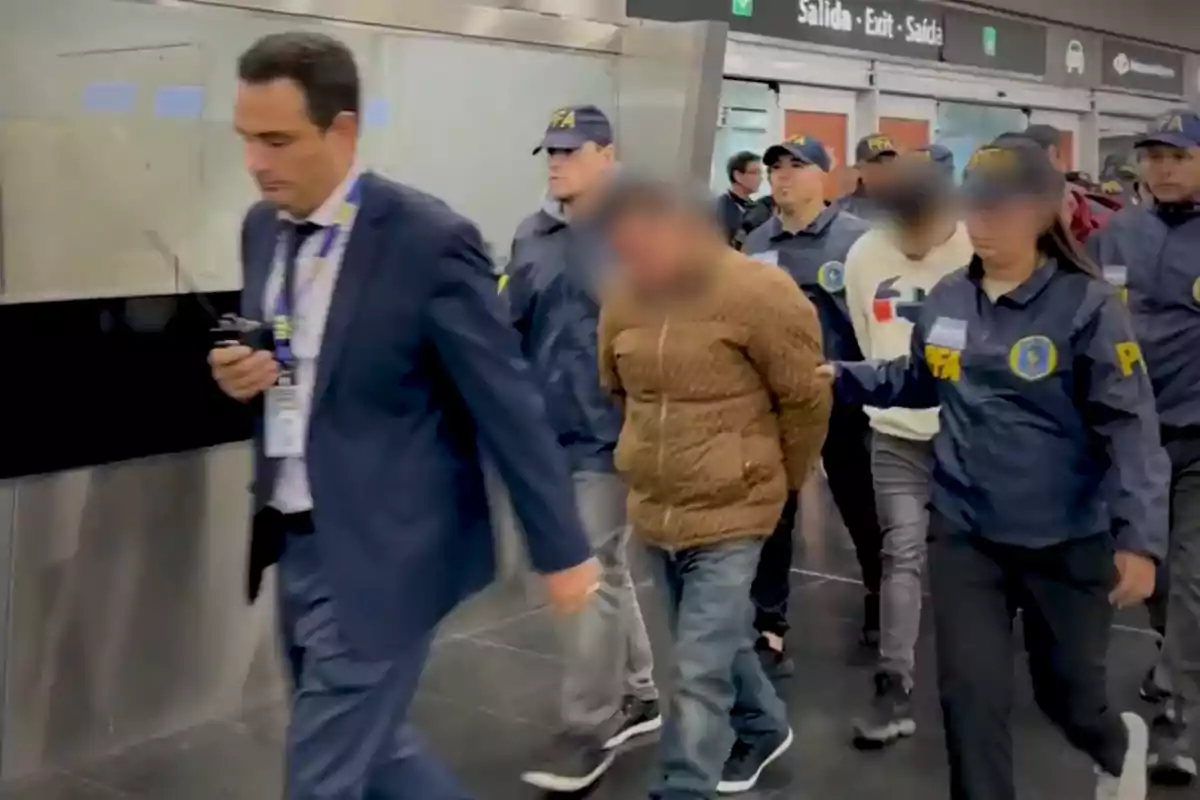 Un grupo de agentes escolta a dos personas esposadas en el interior de un edificio mientras un hombre con traje camina delante de ellos.